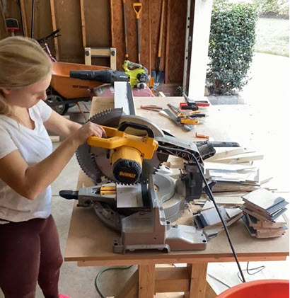 Blonde woman using a miter saw to cut a piece of shiplap for a DIY Shiplap Fireplace.